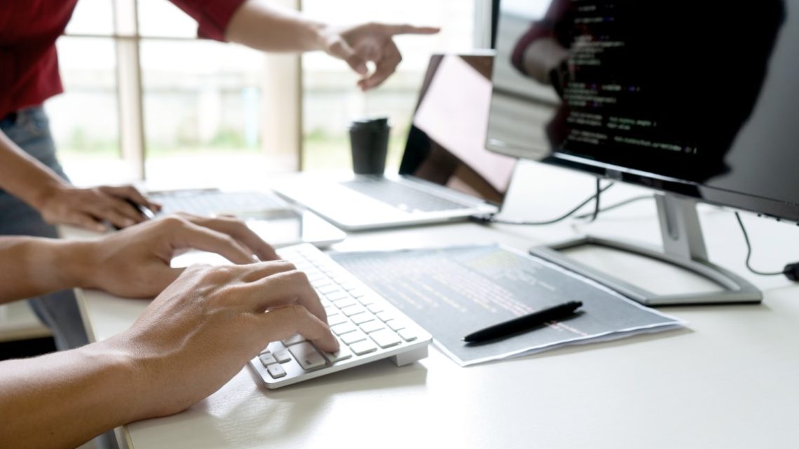 Man pointing towards a computer screen with another person typing website code as part of Website maintenance